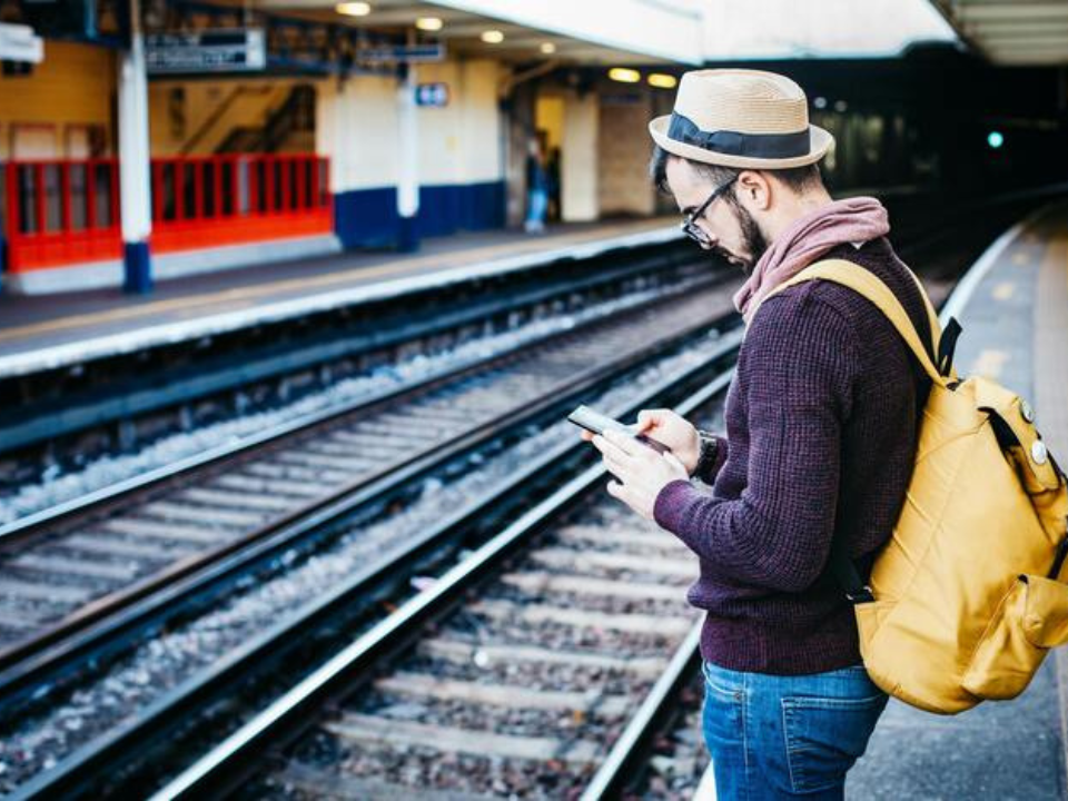 Ein Mann mit gelbem Rucksack wartet bei Gleisen und blickt auf sein Handy