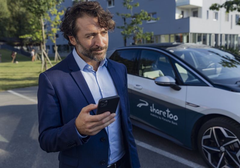 Mann mit dunkelblauem Sakko hat ein Smartphone in der Hand und steht vor einem sharetoo-Fahrzeug