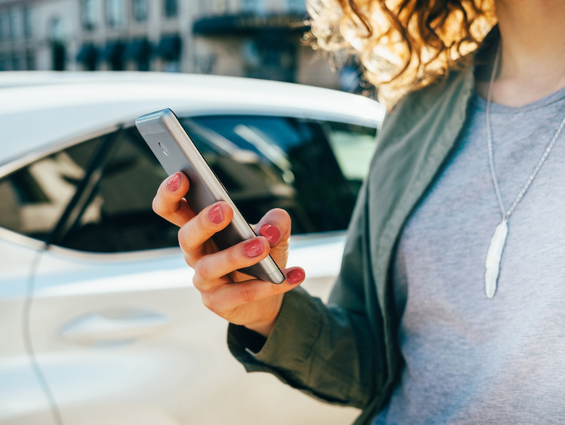 Eine Frau steht vor einem weißen Auto und benutzt ihr Smartphone.